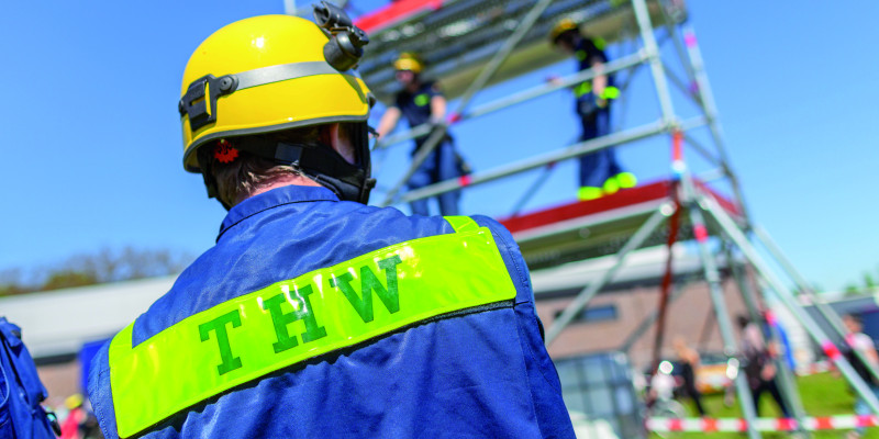 The picture shows a THW member from behind; he is in uniform and wears a helmet. It is possible to make out a scaffold with other THW members standing on it.