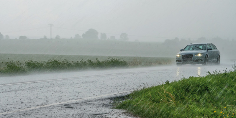 Das Bild zeigt eine mit Wasser bedeckte Straße, auf der ein Auto fährt. Dieses hat Scheinwerfer und Scheibenwischer angeschaltet. Es regnet stark.