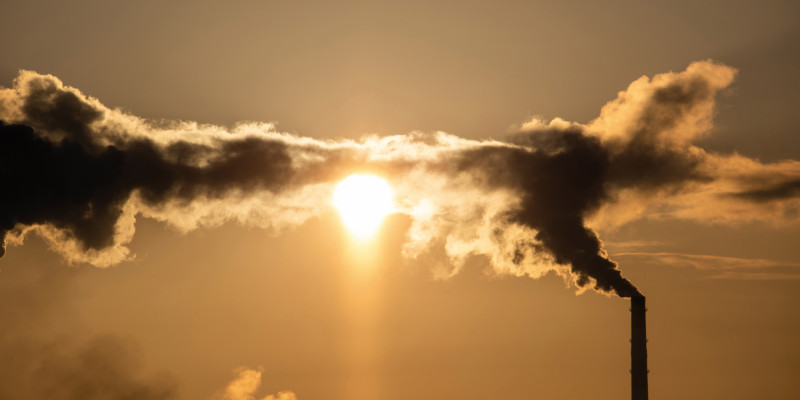 Das Bild zeigt einen qualmenden Industrieschornstein. Die Sonne scheint durch die sich bildenden Dampfwolken und taucht das Bild in warme Braun- und Orangetöne.