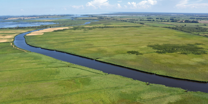 Das Bild zeigt einen Fluss, der durch eine flache Landschaft aus Wiesen und Landwirtschaftsflächen fließt. Im Hintergrund ist das Meer zu sehen.