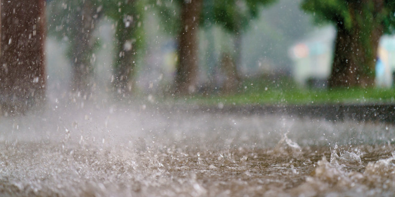 Das Bild zeigt einen Weg oder eine Straße in einem Starkregenereignis. Auf dem Weg steht das Wasser. Die fallenden Regentropfen verursachen vom Boden aufspritzendes Wasser. Im Hintergrund ist verschwommen eine Baumreihe zu erkennen.