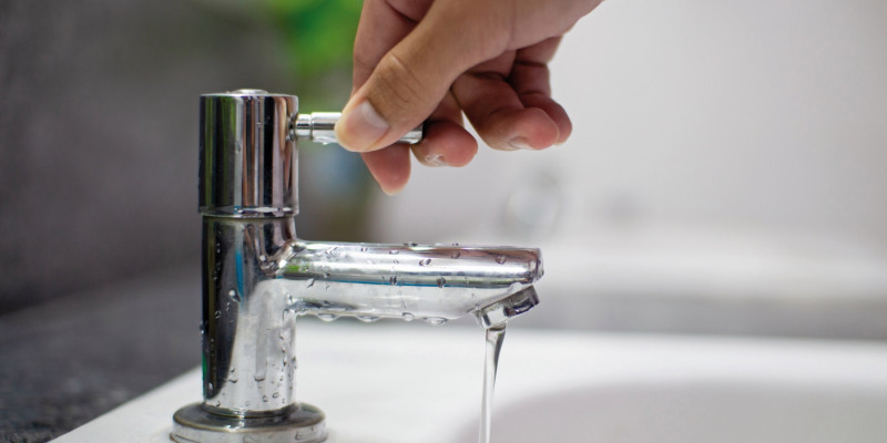 Das Bild zeigt eine Hand am Griff eines Wasserhahns, aus dem ein schwacher Wasserstrahl in ein Waschbecken fließt.