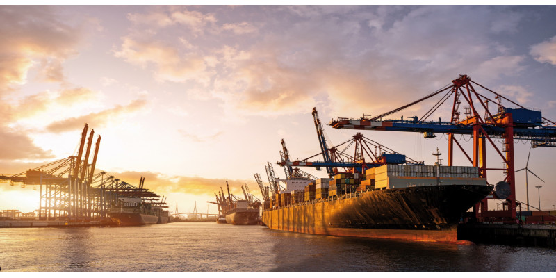 Das Bild zeigt einen großen Containerhafen bei tiefstehender Sonne. Im Vordergrund ist ein beladenes Containerschiff sowie einige Verladekräne zu sehen. Im Hintergrund liegt ein weiteres Containerschiff im Hafen angedockt. 