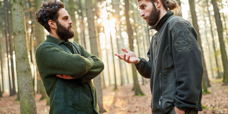 Das Bild zeigt zwei junge Förster, die in einem lichten Wald diskutieren. Der Mann links im Bild verschränkt die Arme, während der andere mit seiner Hand gestikuliert. Im Hintergrund dringen Strahlen der tiefstehenden Sonne in den Wald.