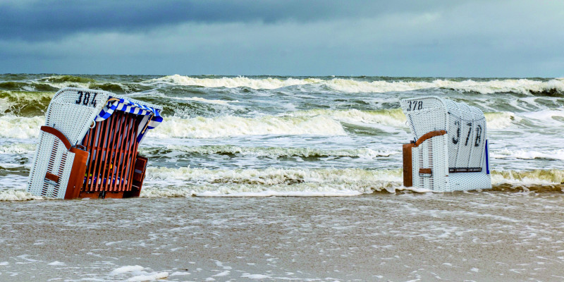 Das Bild zeigt einen Sandstrand an der Ostsee. Ein Strandkorb am Strand wird teilweise vom Wasser umspült. Im Hintergrund sind das Meer mit starkem Wellengang und dunkle Gewitterwolken zu sehen.