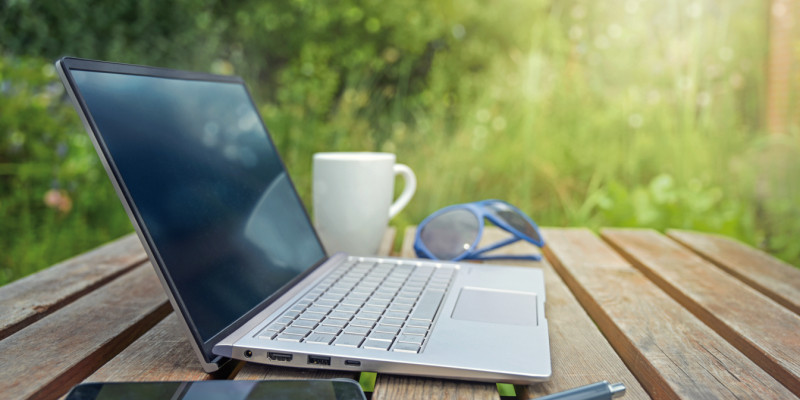 Das Bild zeigt einen Holztisch, auf dem sich ein Laptop, ein Smartphone, eine Kaffeetasse und eine Sonnenbrille befinden. Im Hintergrund sind Sträucher zu sehen. 