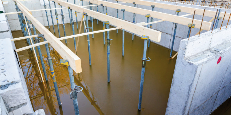 Das Bild zeigt eine Baugrube, in der bereits die Wände eines Kellers stehen. Stahlträger halten Holzbalken, die auf Höhe der Kellerdecke verlaufen. Die Baugrube ist am Boden mit braunem, schlammigen Wasser gefüllt, in dem sich ein Baum spiegelt.