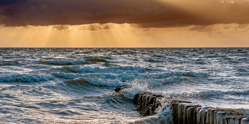 Das Bild zeigt den Ausschnitt eines unruhigen Meeres mit Wellen, die an aus dem Wasser herausragenden Pfählen brechen. Der Himmel ist orange gefärbt und von dunklen Wolken verhangen. Stellenweise treten Sonnenstrahlen zwischen den Wolken hervor.