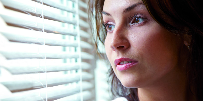 The picture shows a woman’s head and shoulders while she looks sceptically out of a window. The window is part-shaded by an indoor blind.