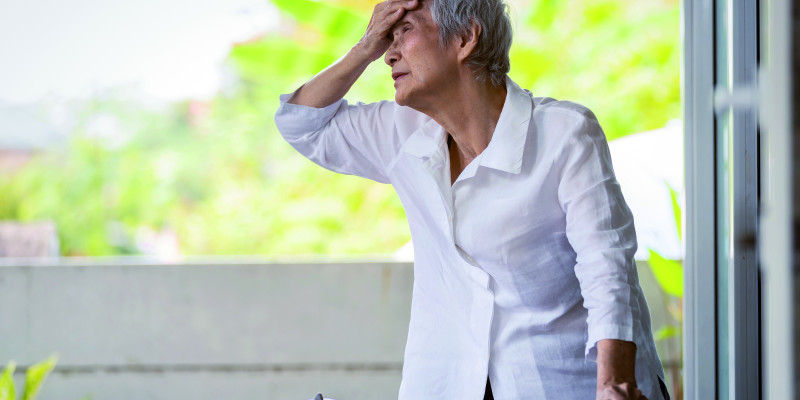 The picture shows an elderly woman wearing a white blouse. The lady is holding on to a rollator with one hand, while she is holding the back of her left hand to her forehead. In the background, a few fuzzy treetops or bushes are visible in bright light.