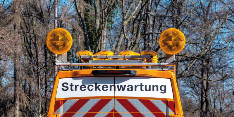 Das Bild zeigt das obere Teil des Hecks eines Fahrzeugs des Straßenbetriebsdienstes. Das Fahrzeug ist orange und seine Hecktüren sind mit dem Schriftzug "Streckenwartung" sowie rot-weiß gestreiften, reflektierenden Aufklebern beklebt. Auf dem Fahrzeug sind Warnlampen montiert.