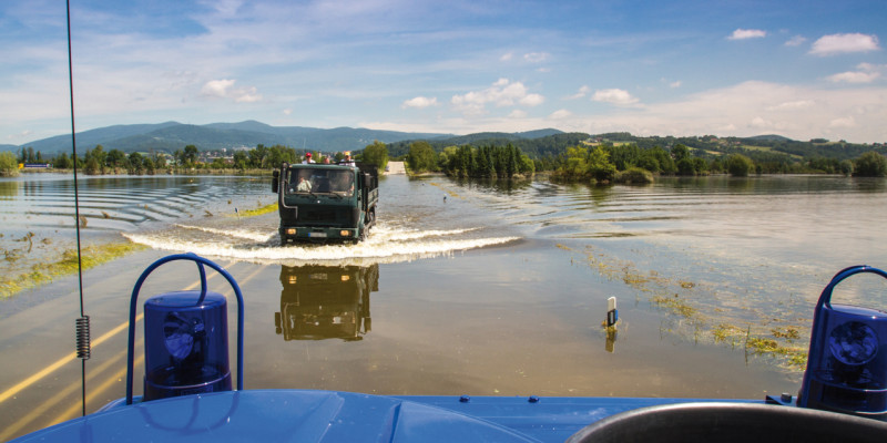 Das Bild zeigt eine von Hochwasser überschwemmte Landschaft. Im Bildzentrum ist eine überschwemmte Straße zu sehen, auf der ein Lastwagen mit Personen auf der Ladefläche fährt. Im Vordergrund des Bildes ist ein Teil eines blauen Einsatzfahrzeugs inklusive blauer Rundumleuchten zu sehen, das ebenfalls auf der überschwemmten Straße unterwegs ist.