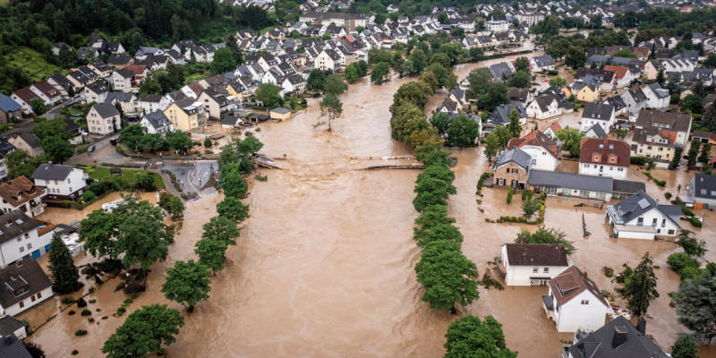 Das Bild zeigt eine Ortschaft mit Ein- und kleinen Mehrfamilienhäusern in einem Tal. Der durch die Ortschaft fließende Fluss ist über die Ufer getreten und überschwemmt die tiefer liegenden Teile der Ortschaft mit braunem Wasser. Der übliche Flusslauf ist durch Baumreihen zu erkennen. Zudem ist eine zerstörte Brücke, die den Fluss querte, sichtbar.