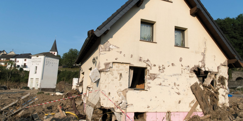 Das Bild zeigt ein stark beschädigtes Einfamilienhaus. Die Außenwand ist an mehreren Stellen durchbrochen worden, Fenster fehlen und der Außenputz ist bis über das Erdgeschoss hinaus löchrig. Um das Haus herum hat sich Schlamm und Schutt gesammelt.