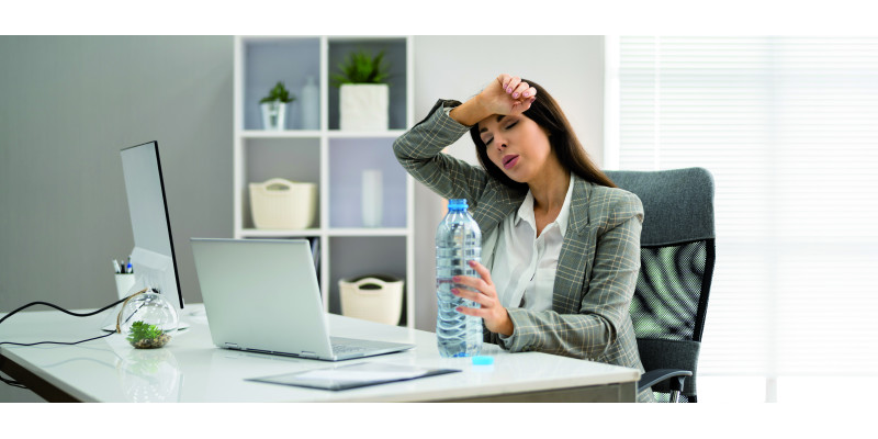 The picture shows a woman wearing a blouse and a blazer, sitting in an office in front of a laptop. In her left hand she is holding an open water bottle while wiping sweat off her brow with her right hand. The woman’s eyes are closed and her facial expression suggests exhaustion.