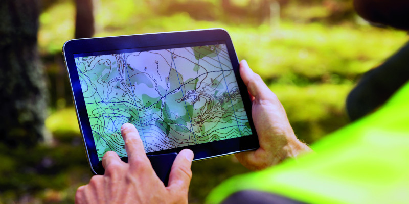 The picture shows hands holding a Tablet with the screen showing a map with contour lines as well as other markings. In the blurred background of the picture it is possible to see that the person holding the Tablet is standing in a wood.