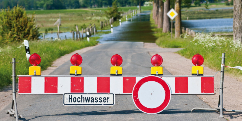 Das Bild zeigt eine durch eine Senke führende überflutete Straße. Im Vordergrund ist sie mit einer Straßensperre abgesperrt. An der Sperre hängt ein Schild mit der Aufschrift "Hochwasser". Rechts und links der Straße sind überflutete Wiesen zu erkennen.