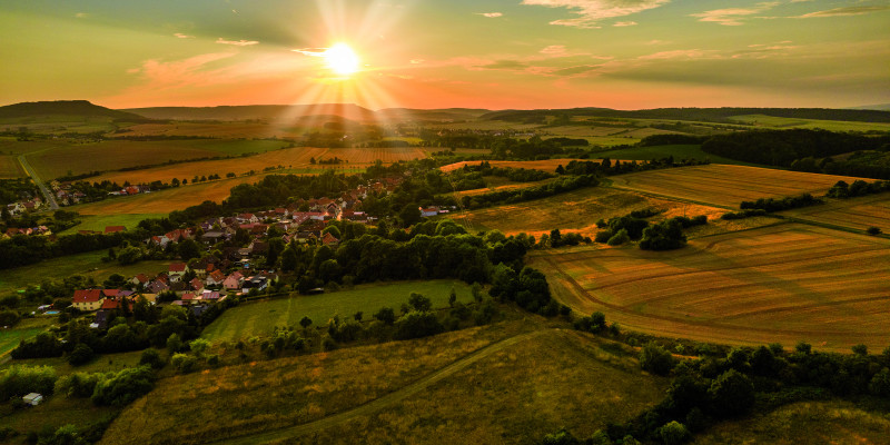 Das Bild zeigt eine leicht hüglige Landschaft, die ackerbaulich geprägt ist. In der Bildmitte ist ein Dorf in die Landschaft eingebettet. Am fast wolkenlosen, zum Teil schon orange gefärbten Himmel steht eine tiefstehende Sonne, die die Landschaft in warmes Licht hüllt.