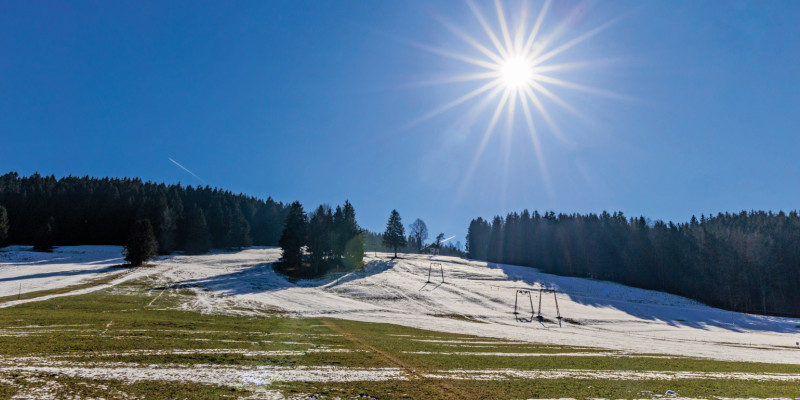 Das Bild zeigt einen Skilift, der auf eine überwiegend mit Nadelbäumen bewaldete Anhöhe führt. Eine dünne Schneedecke liegt auf der Anhöhe. Vor allem im Vordergrund sind aber weite Teile des Bodens ohne Schnee, sodass Rasen und nackter Boden zu erkennen sind. An einem wolkenlosen Himmel steht eine strahlende Sonne.