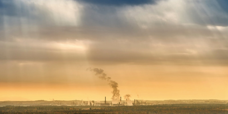 Das Bild zeigt einen bewaldeten Landschaftsausschnitt. In der Ferne ist ein aus mehreren Gebäuden und Anlagen bestehendes Industriegebiet zu sehen. Aus einigen Schloten steigt Rauch empor. Der Himmel ist stark bewölkt. Am Horizont ist er gelblich gefärbt, am oberen Rand des Bildes von dunklen, grau-blauen Wolken verhangen. An wenigen lichteren Stellen dringen Sonnenstrahlen durch das Wolkenband.