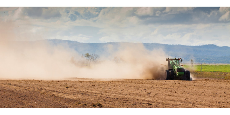 Das Bild zeigt eine trockene Ackerfläche, über die ein Traktor fährt. Aufgrund der Trockenheit wirbelt das Fahrzeug große Staubwolken auf. Im Hintergrund ist eine bewaldete Hügelkette zu erkennen.