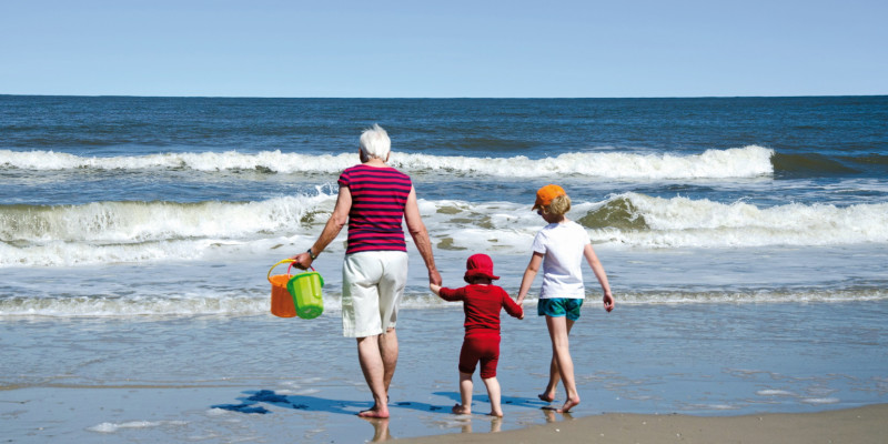 Das Bild zeigt eine ältere Frau, ein Kleinkind und ein Schulkind in Sommerkleidung von hinten. Die drei halten sich an den Händen und laufen am Strand gemeinsam auf die Wellen zu. Die Frau hält zwei Buddel-Eimer in der Hand.