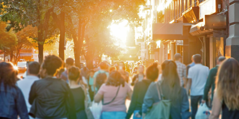 Das Bild zeigt einen städtischen Bürgersteig an einer dicht bebauten Straße im Sommer. Auf dem Bürgersteig sind zahlreiche Personen von hinten zu sehen, einige tragen Einkaufstaschen. Die tiefstehende Sonne hüllt die Szenerie in gelblich-warmes Licht.