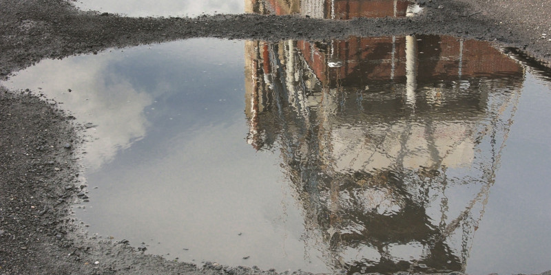 The picture shows two puddles on a gravel surface in which an industrial building is reflected.