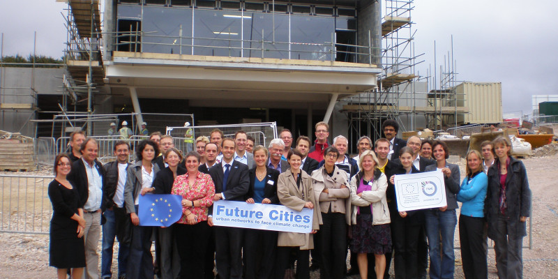 Viele Personen vor einem Haus im Bau, die die EU-Fahne und das Future Cities-Logo hochhalten