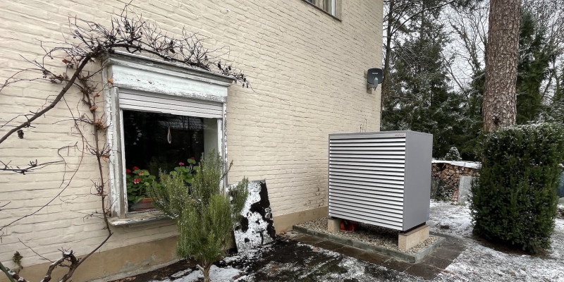 Ein Außengerät einer Wärmepumpe im leicht verschneiten Garten, quer zur beigen, geziegelten Hauswand aufgestellt.