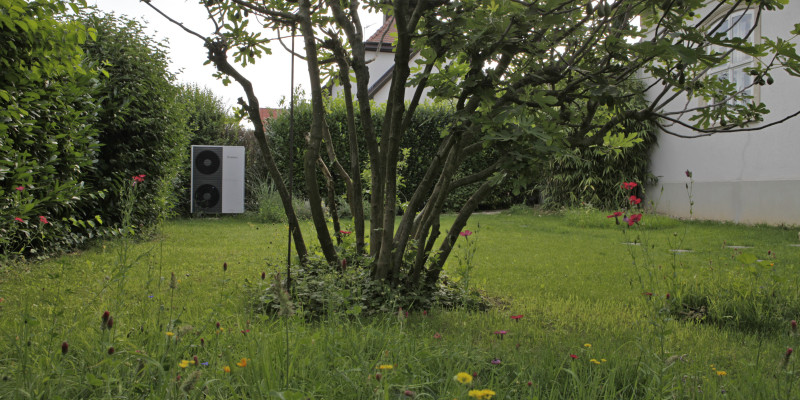 Ansicht des Gartens: auf der Wiese, die eine Hecke säumt, steht in einer Ecke die Außeneinheit der Wärmepumpe; im Vordergrund ein großer Busch.