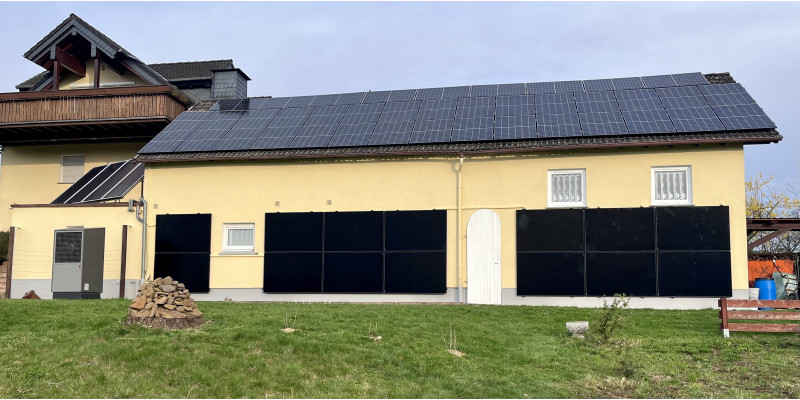 Außenansicht eines Gebäudes auf einer Wiese vor blauem Himmel, inkl Solarthermie und Photovoltaik auf dem Dach