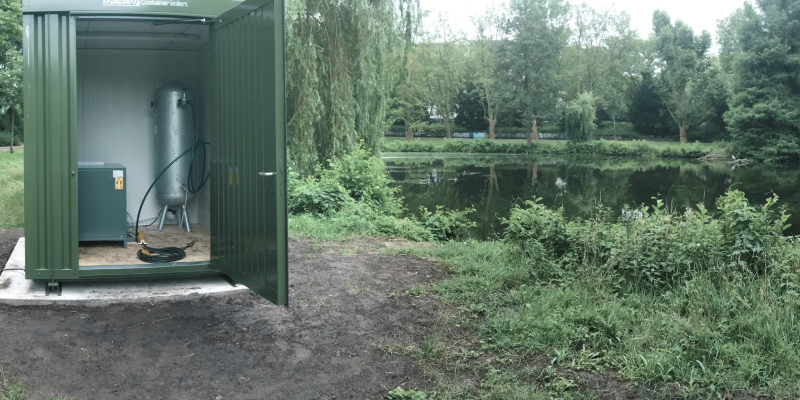 Neben dem Teich steht eine grüne Umhausung - in dem isolierten Container sind Kompressor und Druckluftbehälter untergebracht.