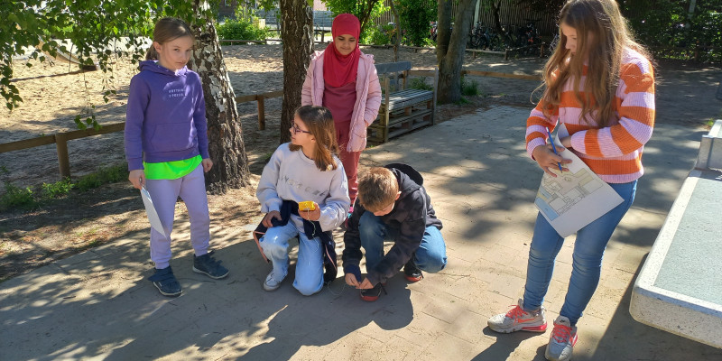 5 Kinder führen Temperaturmessungen auf einem Schulhof durch. Die Sonne scheint. Bäume spenden Schatten. 