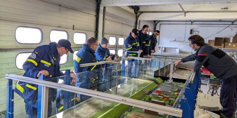 Schulungsteilnehmende blicken in den Schaukasten Hochwassergenese. Die Personen tragen gelb/blaue Arbeitskleidung.