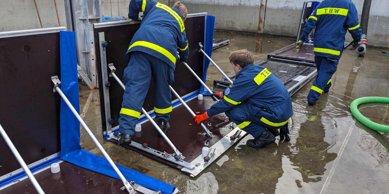  Personen  klappen im praktischen Test System 1 den mobilen Hochwasserschutz auf. Sie tragen blau/gelbe Arbeitskleidung.