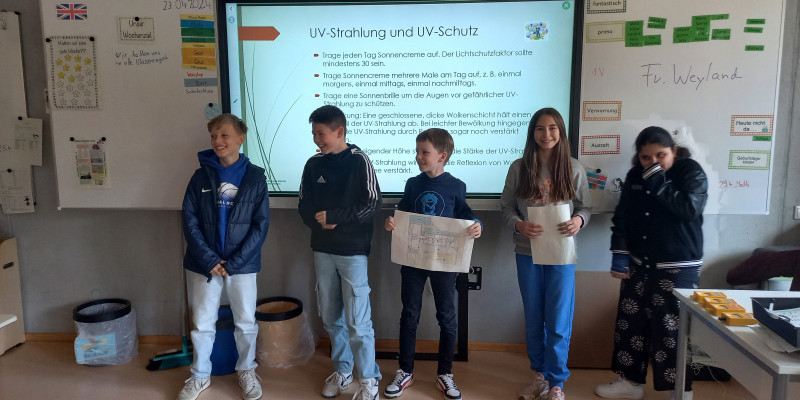 5 Kinder stehen vor einer digitalen Tafel in einem Klassenraum und stellen Ergebnisse ihrer Untersuchungen vor.