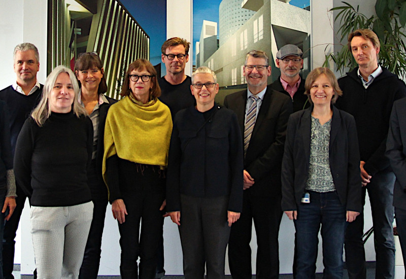 Gruppenfoto mit zwölf Personen in zwei Reihen vor einer Wand mit zwei Architekturbildern.