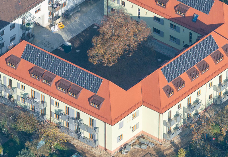 Luftbild eines Wohngebäudes mit drei Flügeln in einem U. Zwei Vollgeschosse und ein Walmdach mit mehreren Gauben und PV-Anlagen. Im Innenhof ein großer Baum, in der Umgebung Kleingärten. Vor der Öffnung des U ein linienförmiger Wohnbau ähnlicher Kubatur.