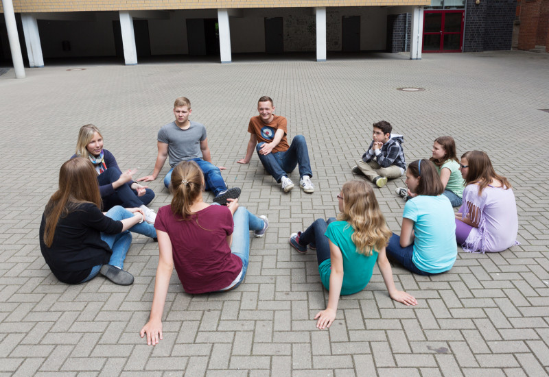 Auf dem Bild sind Jugendliche zusehen, die in einem Kreis auf dem Schulhof sitzen.