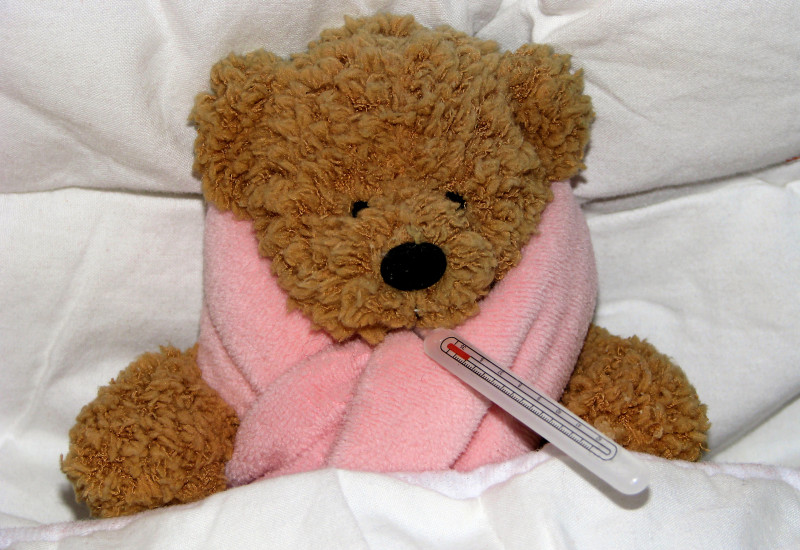 Teddybär liegt mit Schal und Fieberthermometer im Bett