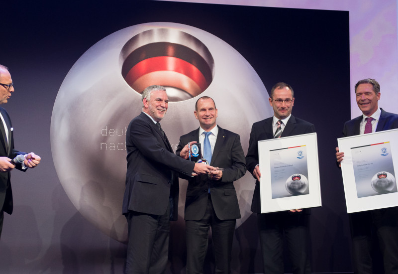 on a stage with a big poster "deutscher nachhaltigkeitspreis 2013" Jochen Flasbarth hands the trophy to a smiling man dressed in a black suit