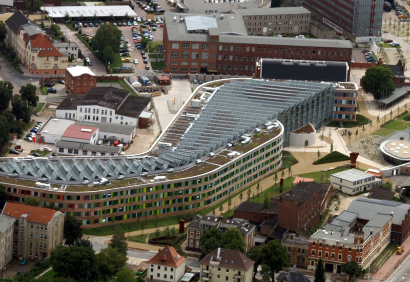  The UBA building in Dessau-Roßlau from above. The building is elongated, with a façade of wood and coloured glass slabs and slanted solar cells covering the entire surface of the flat roof