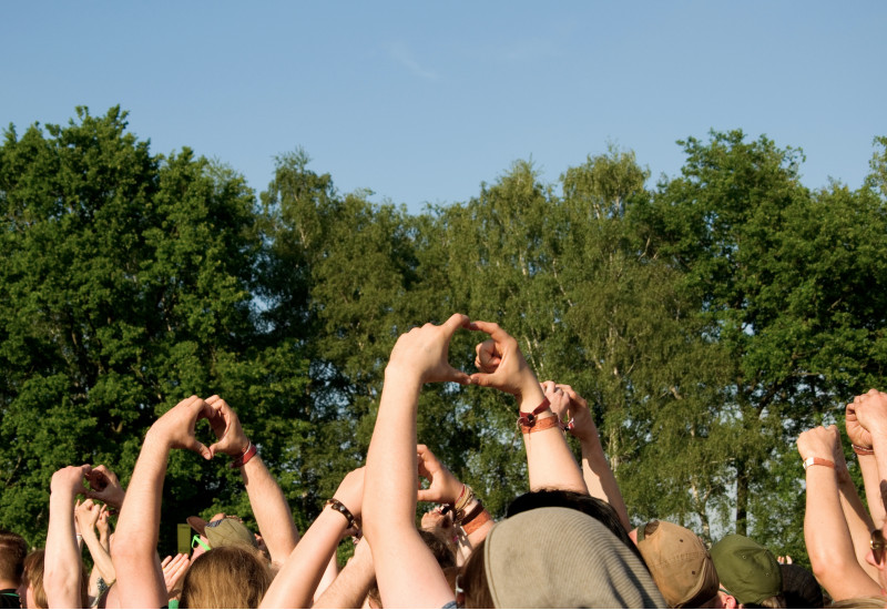 Menschen auf einem Musikfestival im Grünen strecken die Arme in die Höhe und formen mit ihren Händen ein Herz