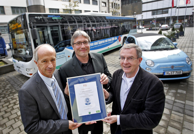 drei Männer halten eine Urkunde mit dem Umweltzeichen "Blauer Engel", im Hintergrund steht ein Linienbus