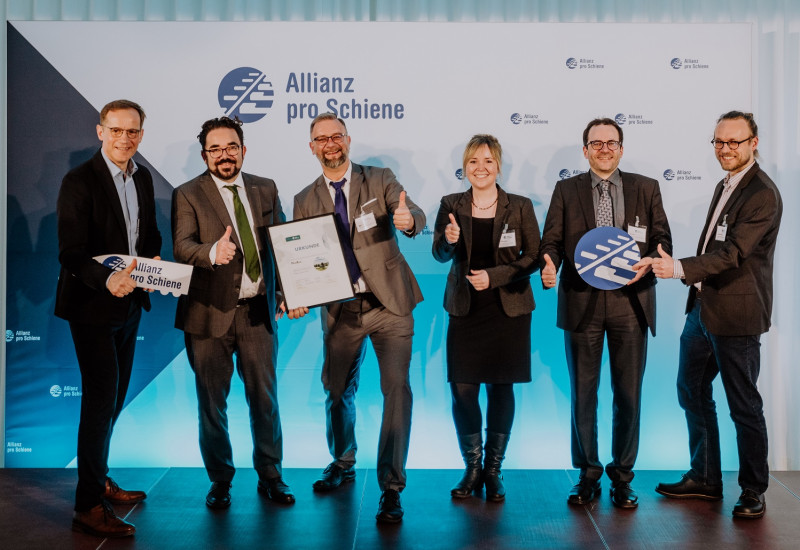 fünf Männer im Anzug und eine Frau im Kostüm stehen lächelnd mit einer Urkunde für ein Gruppenfoto postiert vor einer Stellwand mit dem Logo "Allianz pro Schiene"