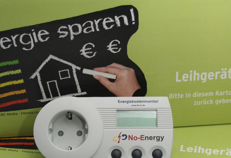 Das Bild zeigt eine hellgrüne Pappkiste mit einem Messgerät für Leerlaufverluste von Elektrogeräten im Vordergrund