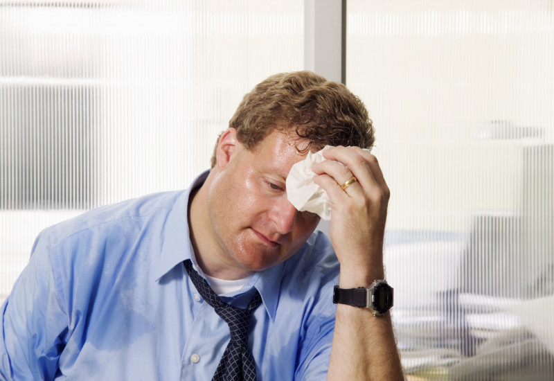 A sweaty man sits in his office in a shirt and tie, wiping his brow.