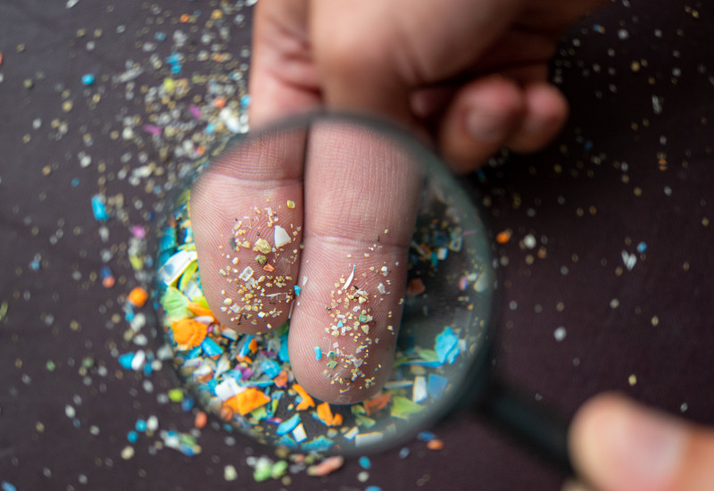 Blick durch eine Lupe auf kleine bunte Mikroplastikteile, die auf zwei Fingern gehalten werden.