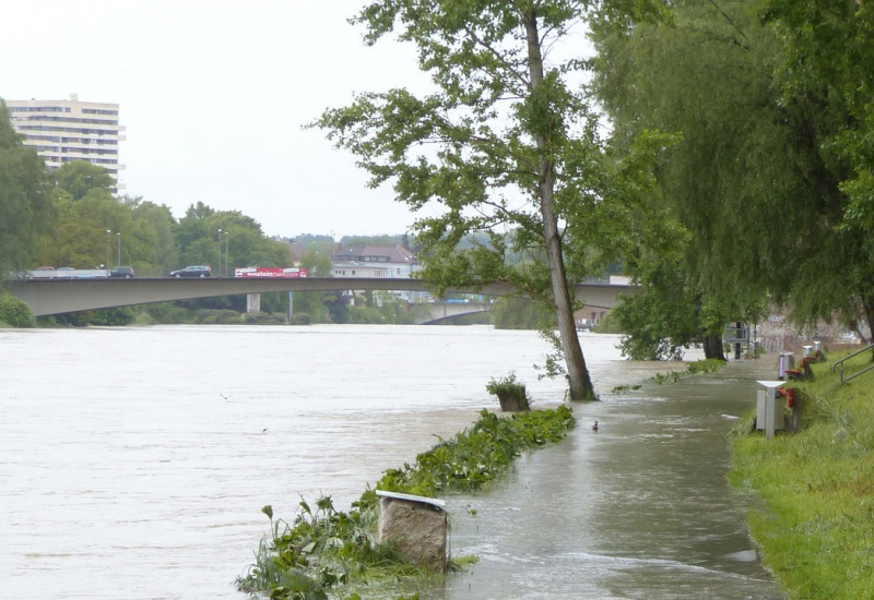 Es ist ein Gehweg zu sehen mit einem Grünstreifen. Dieser ist überschwemmt mit Wasser von dem anliegenden Fluss auf der linken Seite. 